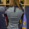 Lifestyle Shot of Youth Female Hockey Player Putting On Protective Shock Doctor Women's Ultra Compression Hockey Long Sleeve Shirt With Integrated Neck Guard (Back View)